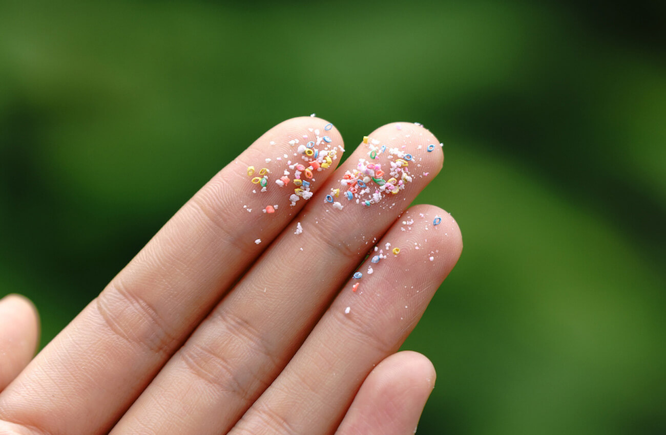 A person holds up their hand in front of a blurred green background - their fingers are covered in microplastics.